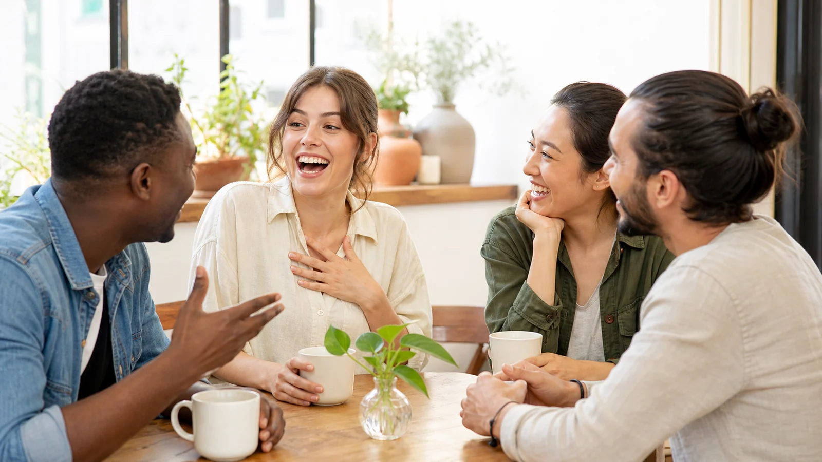 La primera broma que compartimos al instante — A diverse small group at a bright café table shares the exact instant a language learner understands a friendly joke, warm eye contact and laughter together, plain cups, no signage or text.